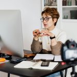 A professional woman engaged in a virtual meeting setup at her home desk, using a computer and camera.